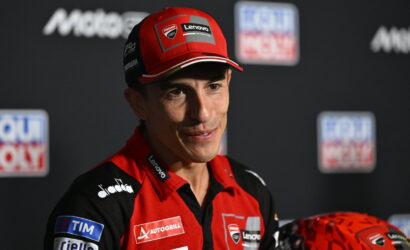 Portrait of a male MotoGP rider in a red and black Ducati Lenovo racing suit at a press event, wearing a red cap with sponsor logos and a red helmet nearby.