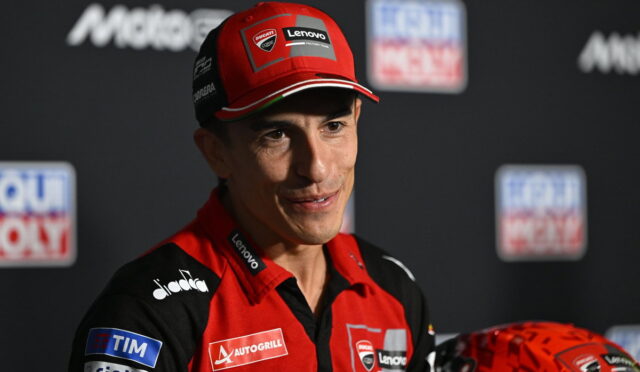 Portrait of a male MotoGP rider in a red and black Ducati Lenovo racing suit at a press event, wearing a red cap with sponsor logos and a red helmet nearby.
