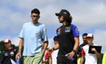 Two men walk together at an outdoor motorsport event; one wears a light blue shirt and green shorts, the other a black and blue Yamaha team shirt with a cap, both holding water bottles.