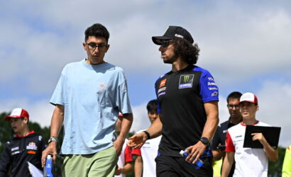 Two men walk together at an outdoor motorsport event; one wears a light blue shirt and green shorts, the other a black and blue Yamaha team shirt with a cap, both holding water bottles.