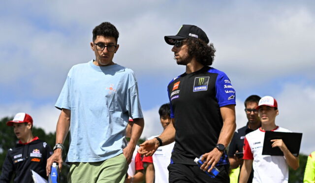 Two men walk together at an outdoor motorsport event; one wears a light blue shirt and green shorts, the other a black and blue Yamaha team shirt with a cap, both holding water bottles.