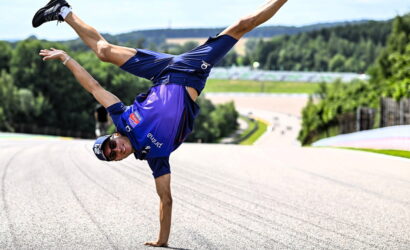 Athlete in blue sportswear doing a one-handed handstand on a race track, sunglasses on, with trees and hills in the background.