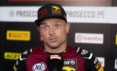 Race car driver in a burgundy racing suit and black cap speaks into a microphone during a post-race interview.
