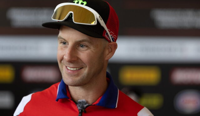 Smiling man in a red jacket with a cap and ski goggles resting on his cap, facing slightly left in an interview setting.