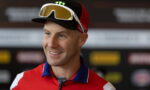 Smiling man in a red athletic jersey with ski goggles resting on a black cap, indoors at a press event.