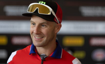 Smiling man in a red athletic jersey with ski goggles resting on a black cap, indoors at a press event.