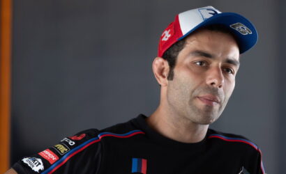 Portrait of a man in a black racing shirt with sponsor patches, wearing a red-white-blue cap, looking off to the side.