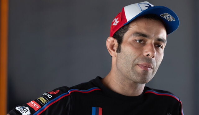 Portrait of a man in a black racing shirt with sponsor patches, wearing a red-white-blue cap, looking off to the side.