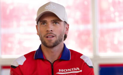 Male race team member in a red Honda Racing jacket and white cap, speaking indoors.