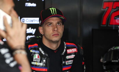 MotoGP rider in a black racing suit with sponsor patches sits in a garage, wearing a Monster Energy cap, looking pensively upward.