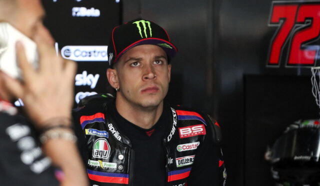 MotoGP rider in a black racing suit with sponsor patches sits in a garage, wearing a Monster Energy cap, looking pensively upward.