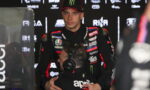 MotoGP rider in a black/red Aprilia racing suit stands in a pit area while being hugged by a team member wearing a headset and sponsor logos around them.