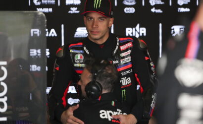 MotoGP rider in a black/red Aprilia racing suit stands in a pit area while being hugged by a team member wearing a headset and sponsor logos around them.