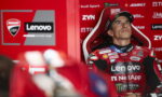 MotoGP rider in a red Lenovo racing suit seated in a red pit area, looking upward.