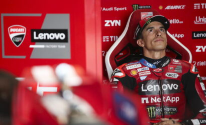 MotoGP rider in a red Lenovo racing suit seated in a red pit area, looking upward.