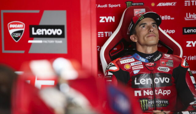 MotoGP rider in a red Lenovo racing suit seated in a red pit area, looking upward.