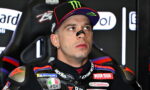 Motorcycle racer in a black cap with Monster Energy logo and a sponsor-filled racing suit sits in a paddock, looking upward thoughtfully.