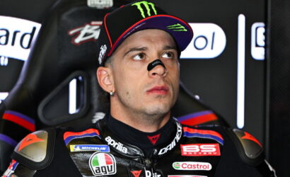 Motorcycle racer in a black cap with Monster Energy logo and a sponsor-filled racing suit sits in a paddock, looking upward thoughtfully.