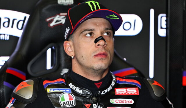 Motorcycle racer in a black cap with Monster Energy logo and a sponsor-filled racing suit sits in a paddock, looking upward thoughtfully.
