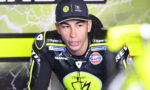 MotoGP rider in black and neon-yellow racing suit and cap, sponsor logos visible in a pit area