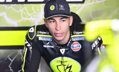 MotoGP rider in black and neon-yellow racing suit and cap, sponsor logos visible in a pit area