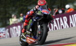 MotoGP rider #20 on a Yamaha race bike leans into a corner, sponsor decals visible on the bike and suit