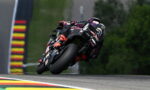 Motorcycle racer on a black sportbike leaning into a left turn on a race track, sponsor logos visible.