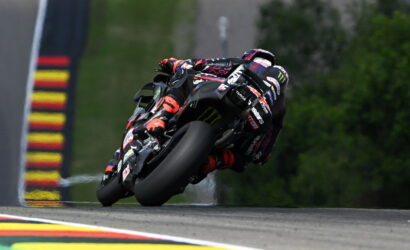 Motorcycle racer on a black sportbike leaning into a left turn on a race track, sponsor logos visible.