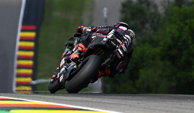 Motorcycle racer on a black sportbike leaning into a left turn on a race track, sponsor logos visible.