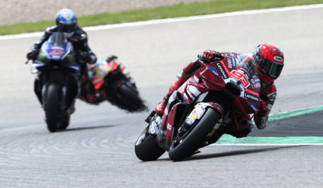 Two motorcycle racers lean into a sharp turn on a racetrack, the red bike leading while a blue-clad rider follows behind.