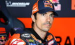 Professional motorcycle racer in racing gear with a Red Bull KTM cap, seated in a press area, looking focused ahead.