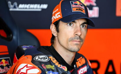 Professional motorcycle racer in racing gear with a Red Bull KTM cap, seated in a press area, looking focused ahead.