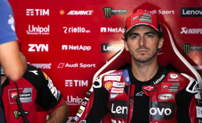 MotoGP rider in red-and-black racing suit sitting in a red sponsor backdrop, with Lenovo and TIM logos visible, looking at the camera.