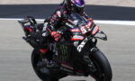 Motorcycle racer in black leathers leaning into a corner on a race track with sponsor decals visible on bike and suit.