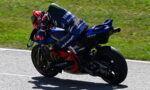 MotoGP rider leans into a left turn on a blue Yamaha race bike with Monster Energy livery on a sunny racetrack edge.
