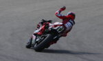 Rider in red racing gear leaning into a turn on a red sport motorcycle on a race track, with sponsor logos visible.