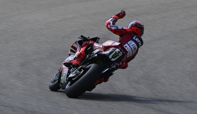 Rider in red racing gear leaning into a turn on a red sport motorcycle on a race track, with sponsor logos visible.