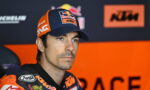 Professional motorcycle racer in KTM/Red Bull gear, wearing a blue cap, seated in the pit area with an orange backdrop.