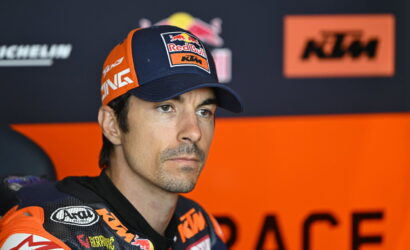 Professional motorcycle racer in KTM/Red Bull gear, wearing a blue cap, seated in the pit area with an orange backdrop.