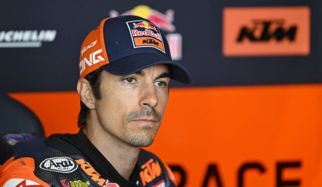 Professional motorcycle racer in KTM/Red Bull gear, wearing a blue cap, seated in the pit area with an orange backdrop.