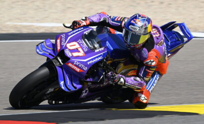 Motorcycle racer in purple livery leaning into a turn on a race track with sponsor logos visible on the bike's bodywork