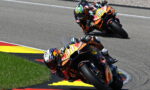 Two motorcycle racers lean into a tight corner on a racetrack, orange Red Bull bikes in a close follow. lAiming their knees close to the asphalt.