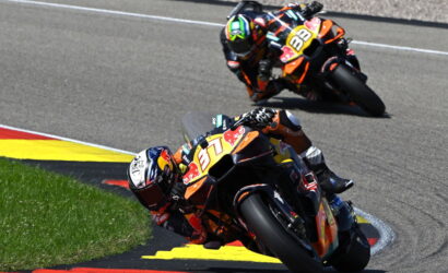 Two motorcycle racers lean into a tight corner on a racetrack, orange Red Bull bikes in a close follow. lAiming their knees close to the asphalt.