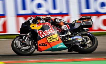 Motorcycle racer on a Red Bull KTM bike leaning into a tight turn on a racetrack, sponsor banners in the background.
