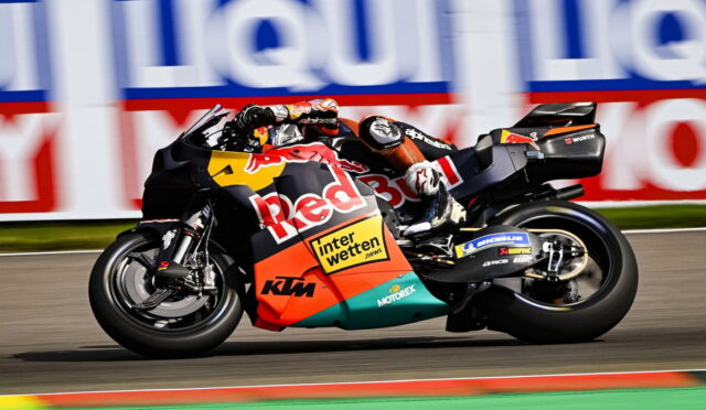 Motorcycle racer on a Red Bull KTM bike leaning into a tight turn on a racetrack, sponsor banners in the background.