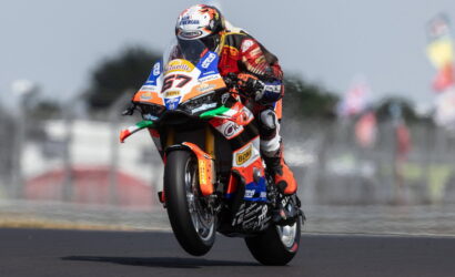 Moto racing rider on an orange, sponsor-labeled bike mid-air during a jump on track, number 67 visible on the front.