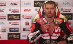 Motorsport rider in red Honda racing suit sits in front of sponsor backdrop during a post-race interview, pointing a small object toward the camera.