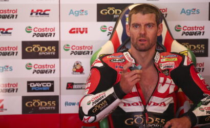 Motorsport rider in red Honda racing suit sits in front of sponsor backdrop during a post-race interview, pointing a small object toward the camera.