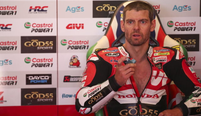 Motorsport rider in red Honda racing suit sits in front of sponsor backdrop during a post-race interview, pointing a small object toward the camera.