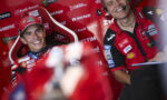 Smiling MotoGP rider in a red racing suit and cap in a sponsor-filled pit with a headset-wearing crew member beside him.